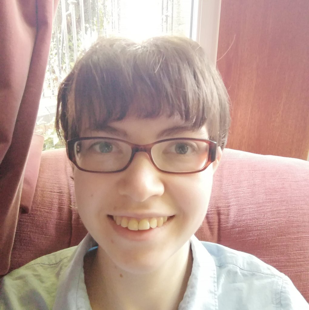 A 26 year old woman with short brown hair and glasses wearing a light blue shirt smiling at the camera whilst sitting on an armchair right in front of a window